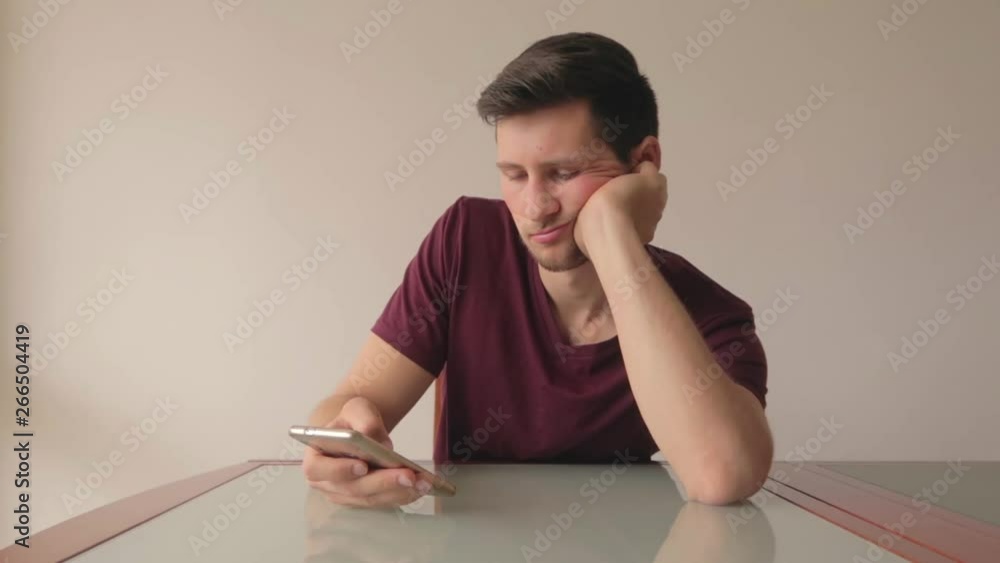 Young man bored looking at phone while sitting at table in minimalist setup, still shot indoor with natural light Young man bored looking at phone while sitting at table in minimalist setup, still shot indoor with natural light