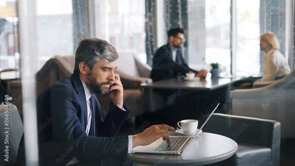 Middle-aged man in suit is making telephone call with smartphone and using laptop working in cafe alone. Business people, communication and gadgets concept. Middle-aged man in suit is making telephone call with smartphone and using laptop working in cafe alone. Business people, communication and gadgets concept.
