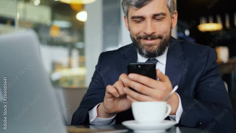Bearded office worker in suit is using smartphone during coffee break touching screen and smiling sitting at table in cafe. Technology and lunch time concept. Bearded office worker in suit is using smartphone during coffee break touching screen and smiling sitting at table in cafe. Technology and lunch time concept.