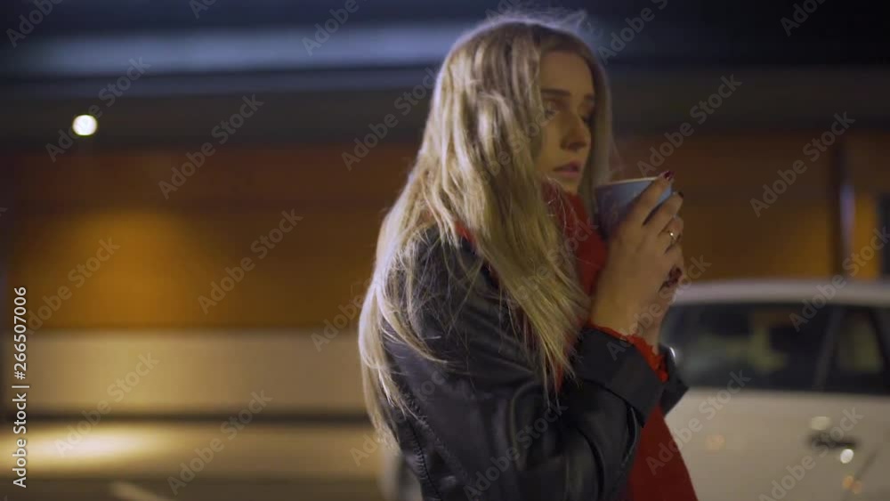 Girl with blonde hair and red scarf is walking on a parking lot while drinking a hot coffee. Girl with blonde hair and red scarf is walking on a parking lot while drinking a hot coffee.