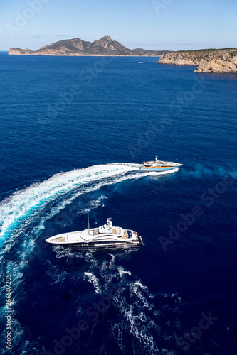Aerial view of luxury big yachts on sea. Big motor yacht running around the luxury yacht creating circles on the water on the background of beautiful mountains. © LaptevArt