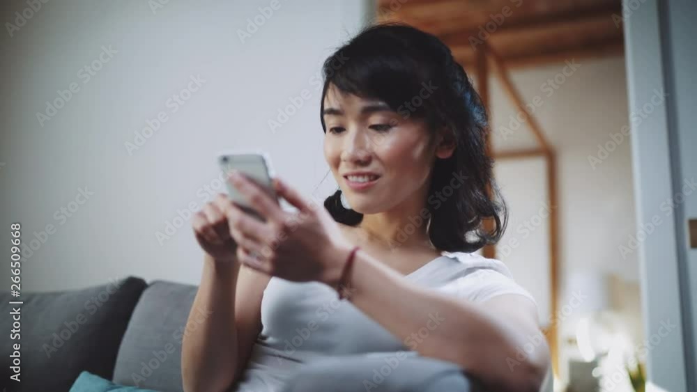 Closeup of beautiful young Asian woman sitting at sofa and using her smartphone at home Closeup of beautiful young Asian woman sitting at sofa and using her smartphone at home