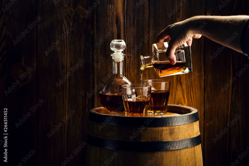 male hand pours brandy into glasses, strong drink in decanters on the barrel, oak barrel in the basement