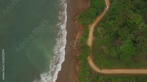 Aerial view of car driving on interesting dirt road between ocean waves and central american jungle. car drives top of scene to bottom and exits right photo