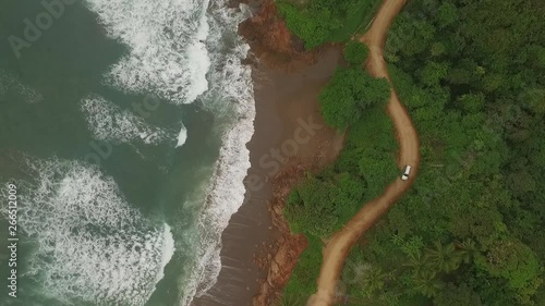Aerial view of car driving on interesting dirt road between ocean waves and central american jungle. camera and car moves middle of scene to bottom photo