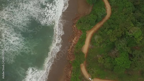 Aerial view of car driving on interesting dirt road between ocean waves and central american jungle. camera and car moves bottom of scene to exit right photo