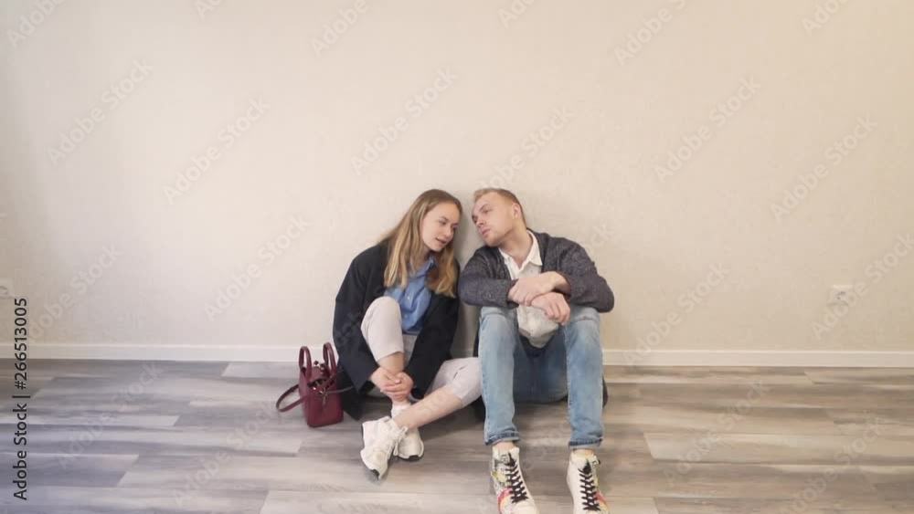 Young Caucasian Married Couple Sitting on Floor In New Empty Finished Real Estate Property Leaning on Each Other and Talk. Light Camera Movement Young Caucasian Married Couple Sitting on Floor In New Empty Finished Real Estate Property Leaning on Each Other and Talk. Light Camera Movement