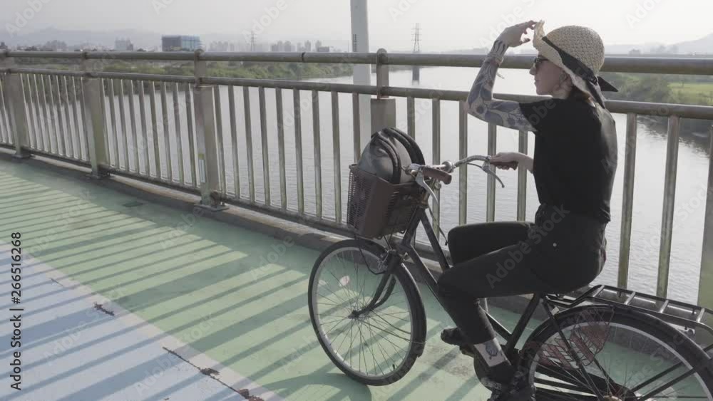 Mirrored shadow cast along tarmac as camera tilts up to girl wearing sunglasses & sunhat riding across transport bridge with views across water & Taipei skyline. Mirrored shadow cast along tarmac as camera tilts up to girl wearing sunglasses & sunhat riding across transport bridge with views across water & Taipei skyline.