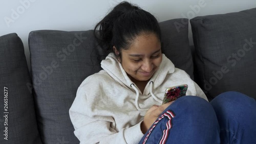 10 year old British Bangladeshi Asian girl, sitting knees up on her mobile smartphone indoors on grey sofa wearing cream hoody photo