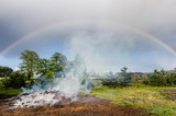 smoke from a large rural bonfire in the countryside slowly rising into the clouds filled sky with a beautiful rainbow during slight rain