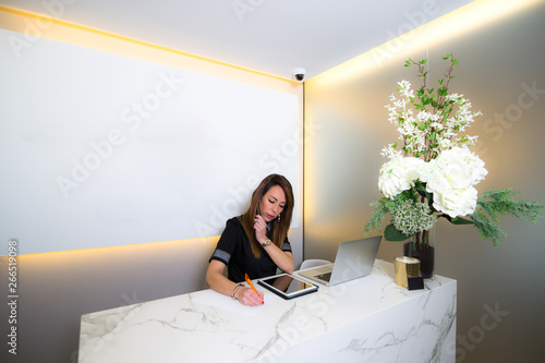 Female practitioner working at the clinic reception. She is taking notes while talking on the phone. Blank screen with copy space in the background.