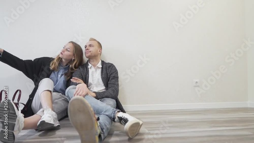 Young Caucasian Married Couple Sitting on Floor in Empty Room of Their New Apartment Bought From Realtor Real Estate Broker. Light Camera Movement photo