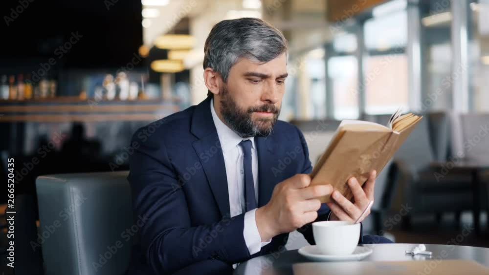 Smart entrepreneur mature bearded man is reading book sitting in cafe alone enjoying interesting story during coffee break. Literature and business people concept. Smart entrepreneur mature bearded man is reading book sitting in cafe alone enjoying interesting story during coffee break. Literature and business people concept.