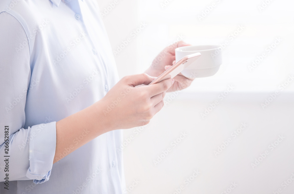 Side view of asia young business woman in blue shirt standing lying on the wall beside window drinking and using smart phone, copy space, close up Side view of asia young business woman in blue shirt standing lying on the wall beside window drinking and using smart phone, copy space, close up