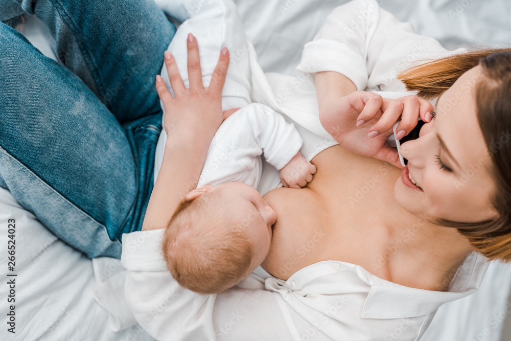 Fototapeta premium smiling woman talking on smartphone while breastfeeding baby at home