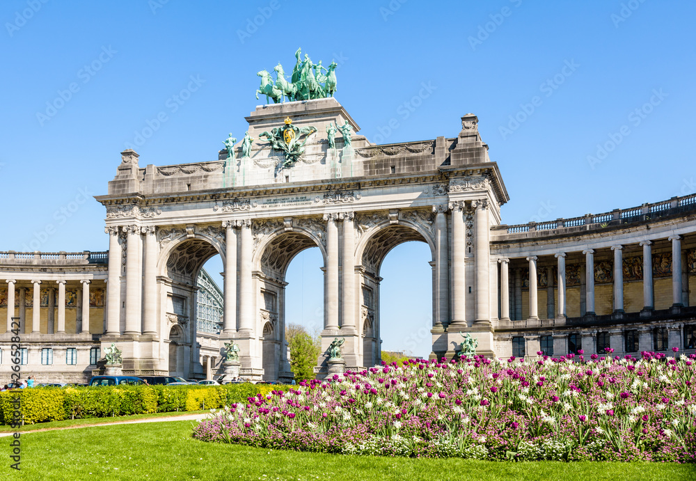Obraz premium The arcade du Cinquantenaire, the triumphal arch erected by king Leopold II in the Cinquantenaire park in Brussels, Belgium, with a flowerbed in full bloom in the foreground against blue sky.