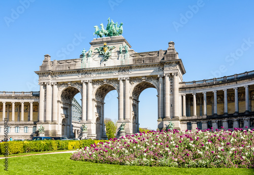 The arcade du Cinquantenaire, the triumphal arch erected by king Leopold II in the Cinquantenaire park in Brussels, Belgium, with a flowerbed in full bloom in the foreground against blue sky.