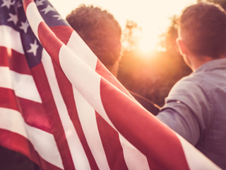 Two friends holding an American Flag against a background of trees and blue sky. View from the back, close-up. National holiday concept