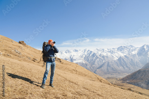 Portrait of a bearded male photographer in sunglasses and a warm jacket with a backpack on his back and a reflex camera in his hands takes pictures against the background of snow-capped mountains