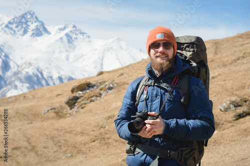 Portrait of smiling bearded male photographer in sunglasses against the background of snow-capped mountains on a sunny day