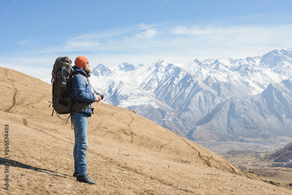 Portrait of a bearded male photographer in sunglasses and a warm jacket with a backpack on his back and a reflex camera in his hands against the background of snow-capped mountains on a sunny day