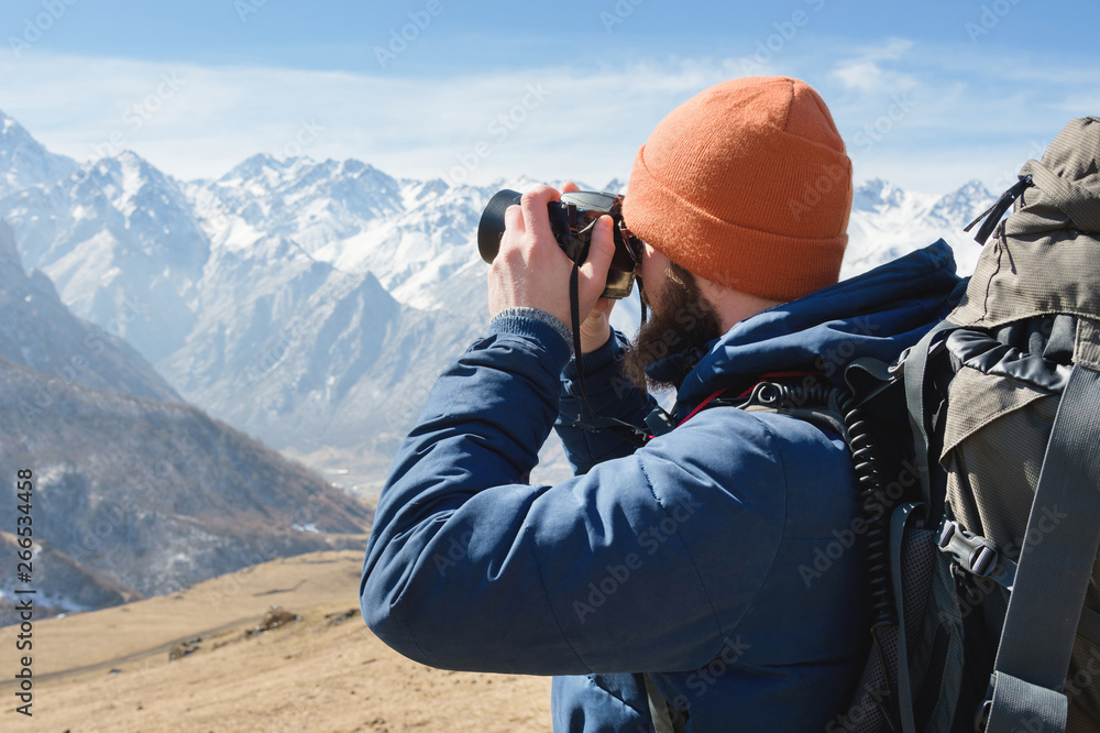 Obraz premium Portrait of a bearded male photographer in sunglasses and a warm jacket with a backpack on his back and a reflex camera in his hands takes pictures against the background of snow-capped mountains