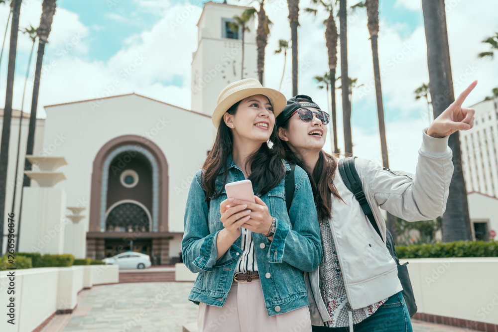 two happy asian woman tourists searching location together with smart phone internet map app and pointing with finger after arrived los angeles walk out union station. joy travelers sightseeing city.