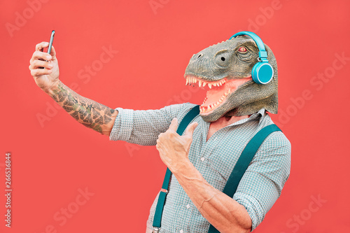 Crazy senior man wearing t-rex mask and taking selfie with mobile smartphone - Hipster older male having fun listening music and dancing outdoor - Absurd, funny and surreal concepts