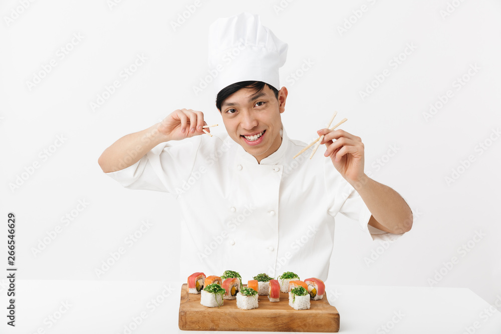 Image of pleased chinese chief man in white cook uniform eating sushi set with chopsticks Image of pleased chinese chief man in white cook uniform eating sushi set with chopsticks