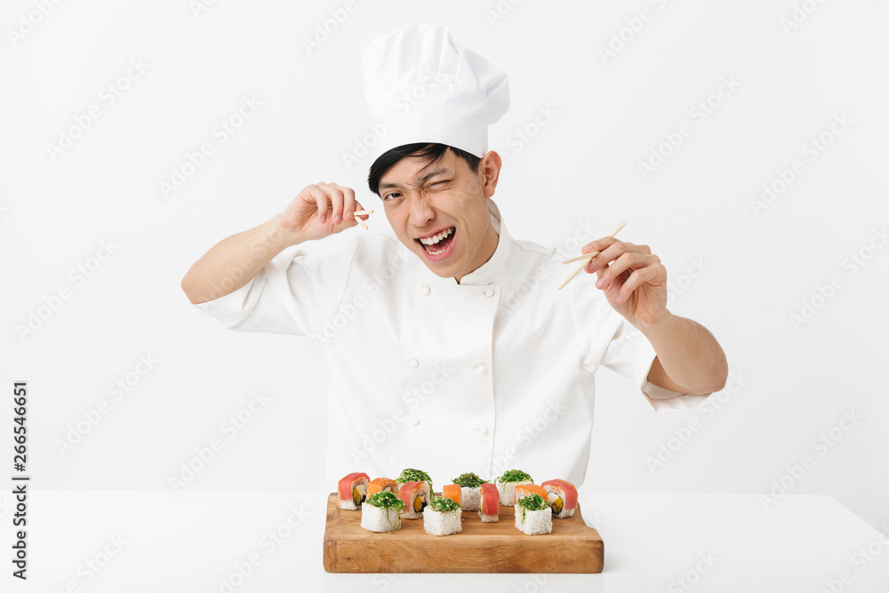 Image of playful asian chief man in white cook uniform eating sushi set with chopsticks Image of playful asian chief man in white cook uniform eating sushi set with chopsticks