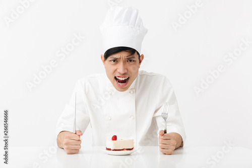 Image of nervous asian chief man in white cook uniform screaming while eating tasty cheesecake
