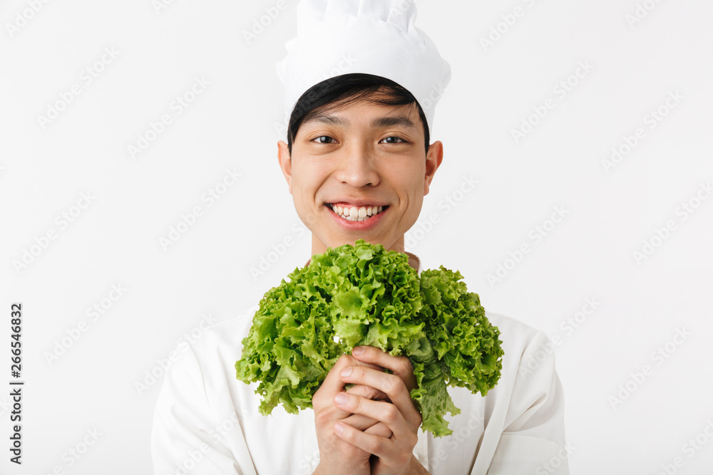 Image of asian joyful chief man in white cook uniform smiling at camera while holding green lettuce salad Image of asian joyful chief man in white cook uniform smiling at camera while holding green lettuce salad
