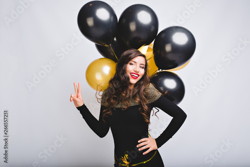 Pretty young woman on white background hold gold and black balloons. Amazing girl with long curly hair, in black elegant dress and yellow crown celebrating new year party, birthday, brightful emotions