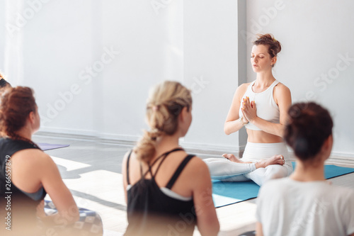 Positive young woman yoga instructor sits in a lotus position next to her unidentified students and teaches them meditation. Group yoga concept