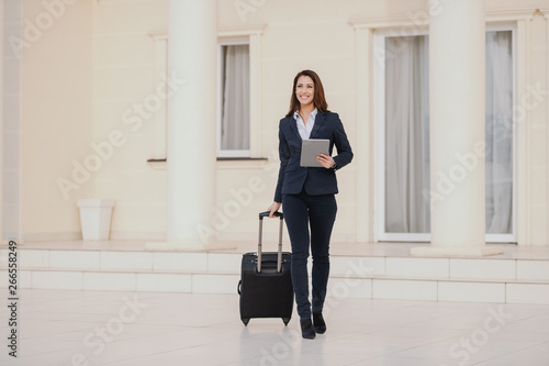 Young smiling brunette dressed smart casual walking, holding luggage and tablet. Business trip concept.