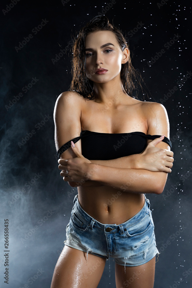 Beautiful leggy and booty athletic fitness girl model, wearing a black bra  ans jeans shorts, with wet oily skin, posing under water drops in  theatrical smoke on a black background Stock Photo