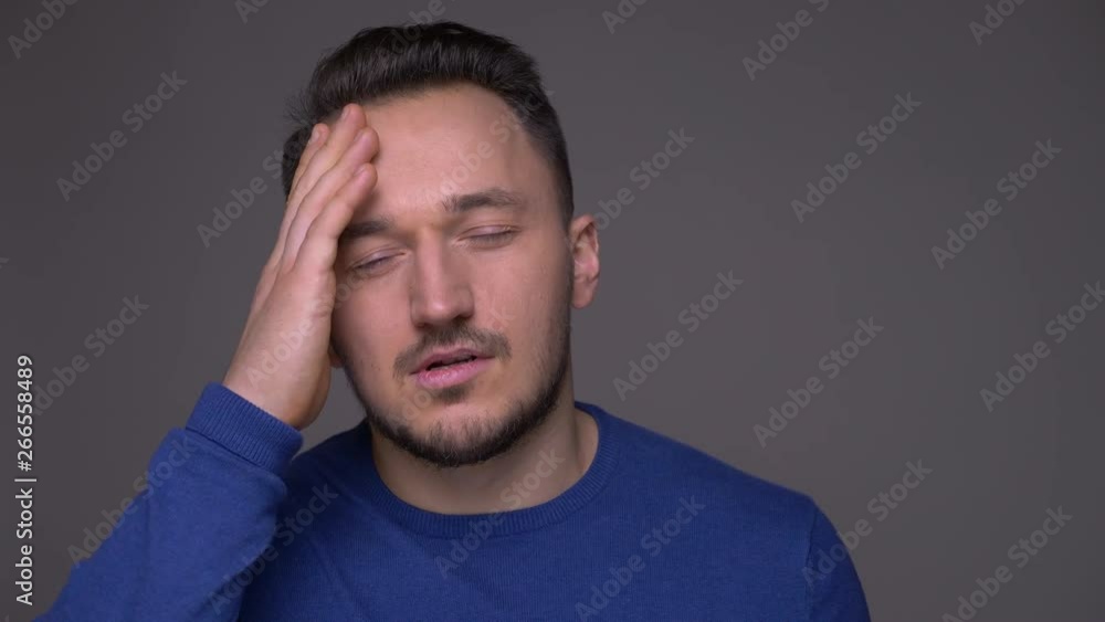 Closeup shoot of young handsome caucasian man having a headache and being exhausted in front of the camera with background isolated on gray Closeup shoot of young handsome caucasian man having a headache and being exhausted in front of the camera with background isolated on gray