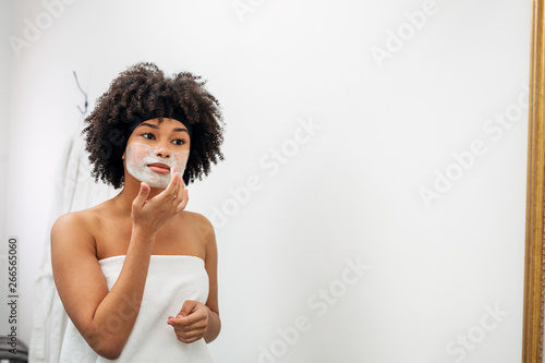 Young woman wrapped in white towel applying natural mask on her face