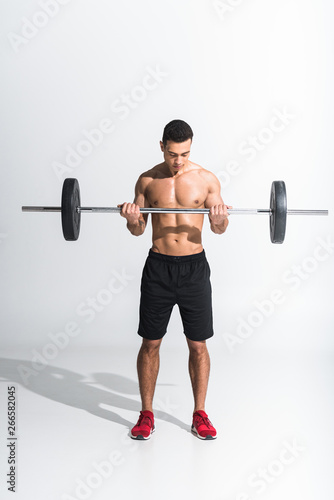 athletic mixed race man in black shorts and red sneakers holding barbell on white