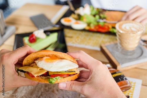 Man holds a burger with egg, chicken, cheese, tomatoes and other vegetables in a restaurant at a joint breakfast with girl. On the background coffee, dessert and main course on a table in a restaurant