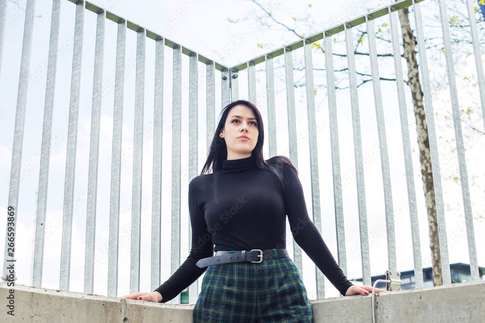 Young woman in black shirt and black hair standing next to fence outdoor in the city, half body, confidence concept, low perspective Young woman in black shirt and black hair standing next to fence outdoor in the city, half body, confidence concept, low perspective