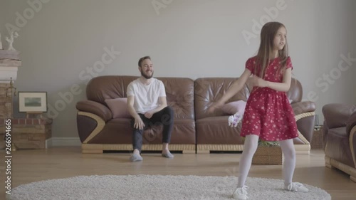 The little funny girl in red dress dancing in the foreground on the fluffy carpet. Bearded father sitting on the background on the leather sofa, clapping his hands and showing big finger