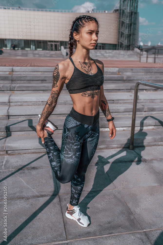 Girl athlete in tattoos, in summer in city, trains fitness, gymnastics  workout. Sportswear leggings top. In the morning before the run. Background  steps. It warms the muscles before exercise. Stock Photo |