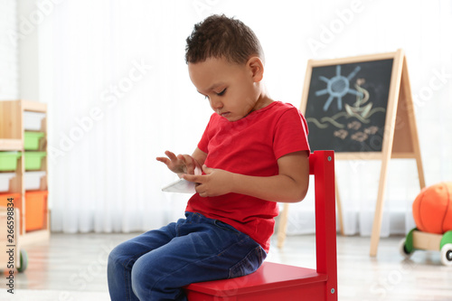 Cute little African-American child playing on mobile phone in kindergarten. Indoor activity