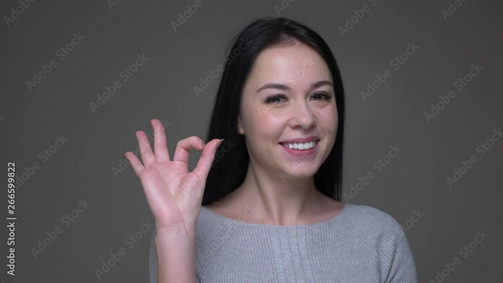 Closeup shoot of young pretty brunette caucasian female showing okay handsign and smiling while looking at camera with background isolated on gray Closeup shoot of young pretty brunette caucasian female showing okay handsign and smiling while looking at camera with background isolated on gray