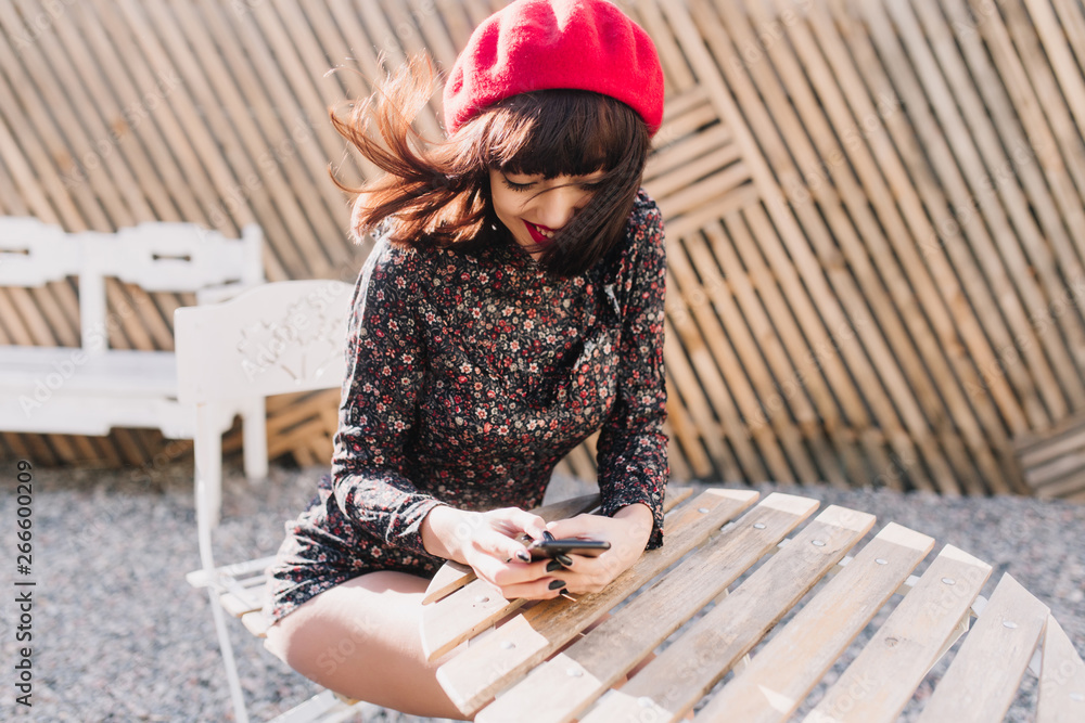 Gorgeous girl with short hairstyle in vintage overall sitting at outdoor cafe with iphone, checking new messages on facebook. Portrait of brunette woman posing at restaurant while wind waving her hair
