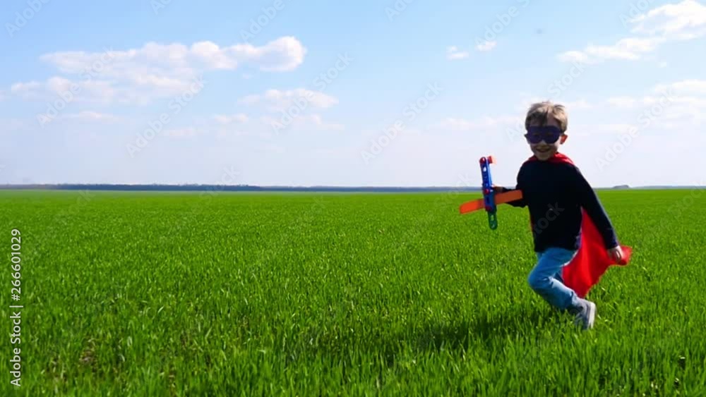 A happy child in a superhero costume runs across the green grass, holding a plane, against a cloudy sky A happy child in a superhero costume runs across the green grass, holding a plane, against a cloudy sky