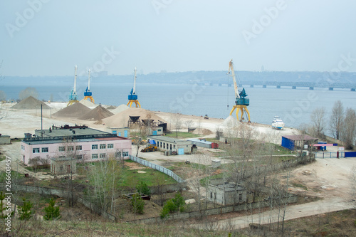 Dock cranes with industrial buildings and big heaps of sand near Volga river in Russia