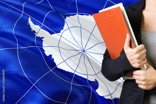 Learning language concept. Young woman standing with the Antarctica flag in the background. Teacher holding books  orange blank book cover.