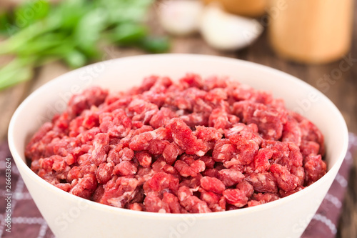 Fresh raw ground or minced beef meat in bowl, seasonings (salt, pepper), garlic and fresh parsley in the back (Selective Focus, Focus one third into the meat)